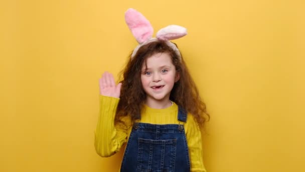 Vidám óvodás kislány nyuszi bolyhos fülekkel nézi a kamerát bájos mosollyal integetve üdvözlő kézzel, ahogy észrevesz valakit, elszigetelve a sárga háttérben. Hello és húsvéti ünnep koncepció