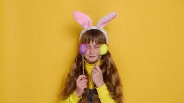 Komik küçük kızın portresi kameraya olumlu bakıyor, gözleri iki Paskalya yumurtasıyla örtüyor, sarı arka planda izole edilmiş tavşancıklar. Mutlu Paskalyalar ve mevsimlik tatil kavramları