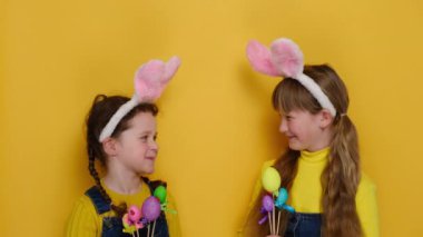 Küçük renkli yumurtalar tutan, dini bayramı simgeleyen, sarı stüdyo arka planında izole edilmiş tavşan kulağı takan iki sevimli kız kardeşin portresi. Paskalya kutlaması kavramı