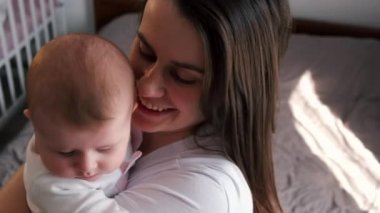 Sevgi dolu genç bir anne kucağında sevimli bir kız bebek kucağında küçük bir çocuğu öpüyor. Sevecen, sevgi dolu bir anne, güzel bir kız çocuğuna sarılıyor ve evde duruyor. Seçici odak