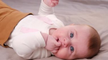 Güzel, beyaz, beyaz, bebek bir kızın, parmakları ağızda, evdeki rahat gri yatakta kameraya bakarak yan görünüm portresi. Çocukluk ve diş çıkarma kavramı