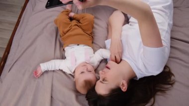 Güzel anne ve bebek kızlarının rahat bir yatakta uzanması, cep telefonu çizgi filmlerini kullanmaları, küçük kızlı annelerinin çevrimiçi arama vlog 'u yapması. Ebeveyn kontrolü, modern teknoloji, genç nesil kavramı