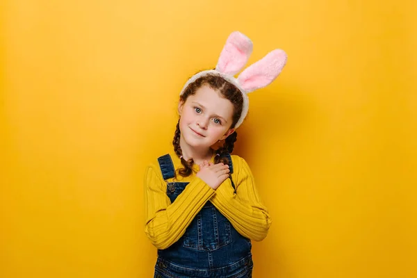 Portrait of smiling preschool girl kid in pink bunny fluffy ears put folded palms on heart, isolated on yellow studio background. Concept easter and sign of sincere kind person, feeling thankful