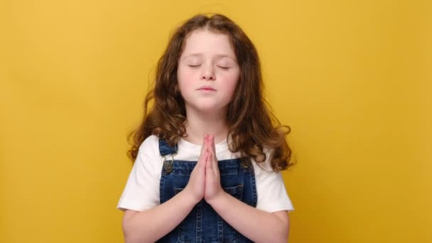 希望のかわいい女の子の子供の祈りの希望で手を保つ幸運を求めると 笑みを浮かべて小さな子供の信者の祈りは 黄色のスタジオの背景に孤立宗教的な迷信的なジェスチャーを行うと信じています 希望の概念 — ストック動画