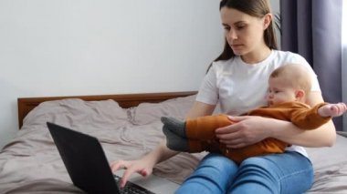 Odaklanmış genç anne girişimci kucağında şirin bir bebek ve dizüstü bilgisayarla evde yatağında araştırma yapıyor. Dürüst girişimci anne çalışıyor ve ebeveynlik yapıyor. İnsanlar internet teknolojisi kavramı
