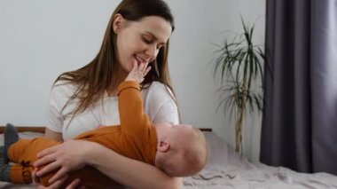Şefkatli bir genç anne, sevimli, komik bir kız çocuğunu kucağına alıp ellerini öpüyor. Sevecen dadı anne evde rahat bir yatakta oturan sevimli bebekle güzel bir anın tadını çıkarıyor.