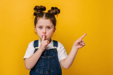 Adorable little girl holding finger on lips symbol of hush gesture of asking to be quiet, isolated over yellow studio background with copy free space for advertisement. Silence or secret concept clipart