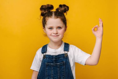 Çekici, küçük bir anaokulu kızı, iyi bir jest yapıyor, sarı stüdyo arka planında fotokopi alanı ile izole edilmiş. Küçük mutlu çocuk portresi fikri kabul ediyor. Anlaşma, onay, onay, tamam konsept