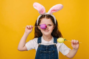 Pembe tavşancık kulaklı küçük kızın yakın portresi göz kapaklarını küçük Paskalya renkli yumurtalarla kaplıyor. Stüdyo sarı arka planda izole edilmiş. Çocukluk yaşam tarzı Mutlu Paskalyalar