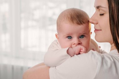 Mutlu, genç, beyaz bir anne yatak odasının penceresinin yanında sevimli bir kız çocuğu tutuyor. Neşeli yetişkin anne, güzel komik çocuk bebekle oynuyor içeride birlikte eğleniyor.