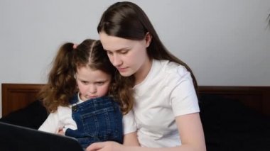 Küçük anne ve küçük kızı önemseyen, dizüstü bilgisayar kullanarak eğlenen, küçük kıza online alışveriş yapmayı öğreten anne, webcam video görüşmesi yapan, bilgisayar ekranına bakan, evde yatakta oturan.