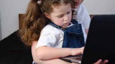 Mutlu genç anne, sevimli küçük kıza ders veriyor evdeki rahat yatakta dizüstü bilgisayarı kullanıyor, gülümsüyor anne ve küçük kız, bilgisayarını tutuyor ve birlikte eğleniyorlar.