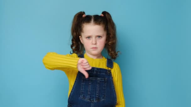 怒り狂った顔の表情小さな女の子がカメラを見て否定的なフィードバックのジェスチャーシンボルを親指ダウンし 青のスタジオの背景に隔離された表示されます 不一致と拒絶の概念 — ストック動画