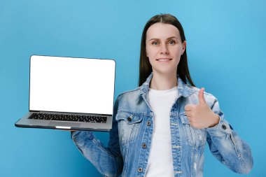 20 'li yaşlarda beyaz bir kadın dizüstü bilgisayarla boş ekran çalışma alanı kullanıyor. El hareketlerini gösteriyor. Kot ceket giymiş, mavi stüdyo arka planında izole edilmiş.