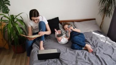 Evde modern aletler kullanan çocuklu bir aile. Gözlüklü genç anne dizüstü bilgisayar ekranına bakarken not alıyor bebek ve küçük kızının yanında oturuyor.