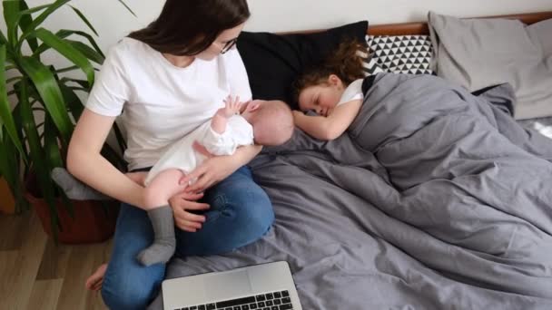 成年妇女戴着眼镜 用笔记本电脑上班 抱着可爱的婴儿 坐在舒适的床上 靠近睡在客厅里的小女儿 年轻的商界妈妈工作和养育子女 — 图库视频影像