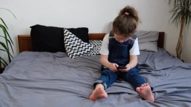 Sevimli küçük anaokulu kızı rahat yatak odasında oturmuş elinde akıllı bir telefonla yeni mobil uygulama kullanarak online video oyunlarına dalmış. Modern teknoloji bağımlılığı olan yeni nesil