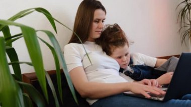 Küçük kızıyla rahat bir yatakta oturup dizüstü bilgisayar seyreden ve evden video konferansı yapan genç bekar bir annenin yan görüntüsü. İki kişilik aile internet mağazasından alışveriş yapıyor.