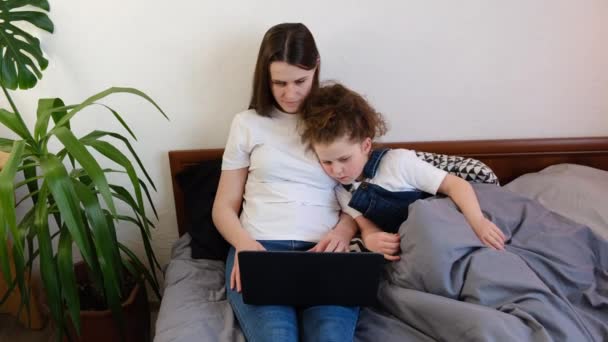 小さな子供の娘がビデオ漫画を見たり ラップトップで一緒にゲームをしたり ベビーシッターや乳母教育の子供の女の子と一緒にコンピュータを使用して自宅でベッドの上に座っています デバイスの使い過ぎの概念 — ストック動画