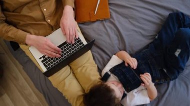 Babanın dizüstü bilgisayarda daktilo kullanışının üst görüntüsü, kızının cep telefonu uygulamalarını kullanarak eğlenmesi. İstemsiz kişisel izolasyon sırasında uzaktan çalışma. Rahat bir çalışma ortamı ve konforlu bir ev.