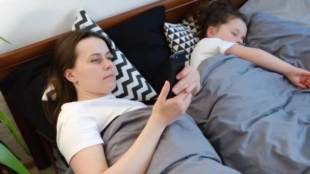 年轻的高加索籍母亲用户手持手机短信躺在熟睡的女儿身边 妈妈在睡前使用智能手机应用程序在手机应用程序中查看社交媒体 — 图库视频影像