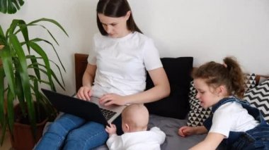 Genç iş anneleri, küçük kızları modern evde vakit geçiriyorlar, tatlı çocuklar rahat bir yatakta uzanıyorlar, odaklanmış anneler bilgisayar kullanımıyla uğraşıyorlar. Konforlu ev aile eğlence aktiviteleri konsepti