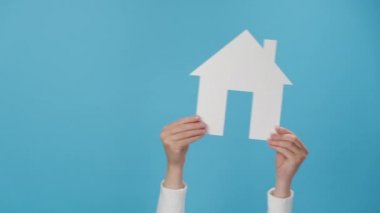 Küçük beyaz kağıt evi tutan kadınların ellerini kapayın, sağa sola sallayın, reklam için fotokopi alanı olan pastel mavisi arka plan duvarı üzerinde izole edilmiş. İpotek ve satış kavramı