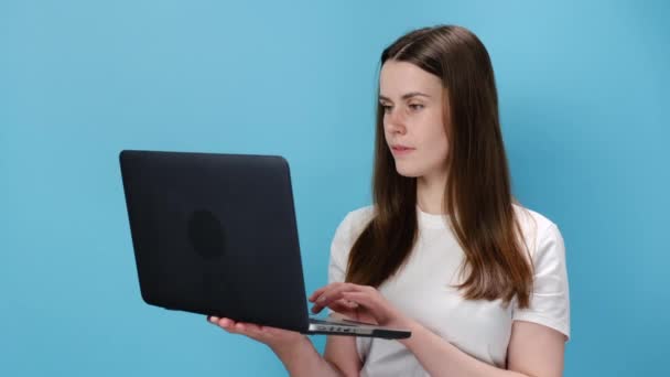 20代の若い白人女性がノートパソコンのコンピュータに入力して親指をジェスチャーのように見せ 白いTシャツを着て 青いスタジオの背景に隔離されています 人々の感情やビジネスコンセプト — ストック動画