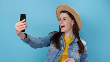Akıllı telefon üfürürken selfie çeken neşeli Avrupalı genç kız hava öpücüğü ver yanağına el koy, kot ceket ve şapka giy, mavi stüdyo arka planında izole ol. Halk yaşam tarzı konsepti