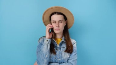 Sinirli, üzgün, beyaz, genç bir kadın akıllı telefonuyla konuşuyor, vıdı vıdı jaja elleri, kot ceket ve şapka giymiş, mavi stüdyo arka plan duvarında soyutlanmış. Halk yaşam tarzı konsepti
