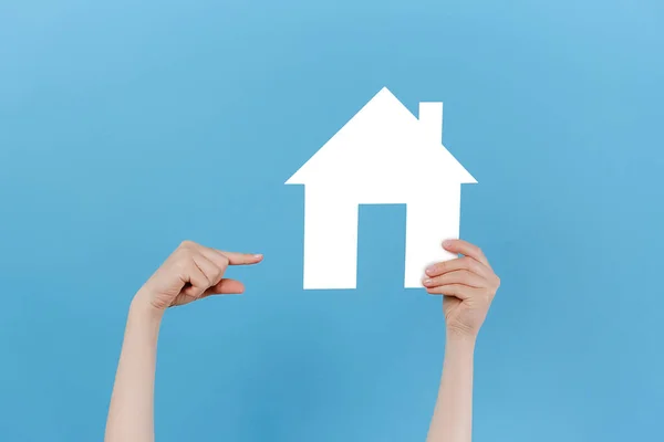Close up of young female hand raising and pointing to small white paper house, posing isolated over blue studio background wall with copy space for advertisement. Property and mortgage concept