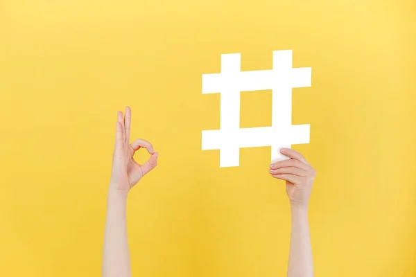 Close up of unrecognizable female holding large big hashtag sign and showing okay gesture, viral web content, internet promotion, isolated on pastel yellow wall. Blogging blog website strategy concept