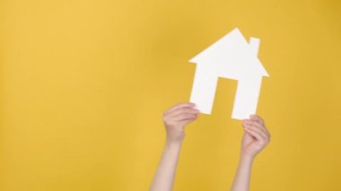 Tanımlanamayan genç bir kadının elinde küçük beyaz bir kağıt ev tutarak, sağa sola sallanarak, reklam için fotokopi çekilen sarı arka plan duvarının üzerinde izole edilmiş bir şekilde. İpotek ve satış kavramı