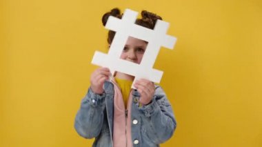Elinde hashtag sembolü olan neşeli, sevimli küçük kız ve kameraya bakarken dişlek gülüşlü, esrar işareti gösteren, sarı arka planda izole edilmiş. Moda sosyal medya gönderileri ve blog yazma kavramı