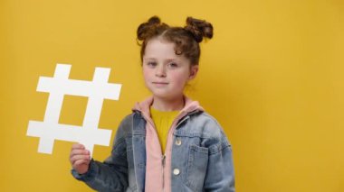 Popüler blog ve trendleri severim. Büyük beyaz hashtag sembolü taşıyan çekici küçük bir kız çocuğu. Başparmak işareti yapıyor ve jest yapıyor. Kot ceket giyiyor, sarı arka planda izole edilmiş.
