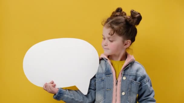 かなりかわいい女の子の子供を保持し 広告のためのコピースペースと黄色のスタジオの背景の壁に隔離されたデニムジャケットに身を包んだ 空白のスピーチバブルを指しています 人々の感情の概念 — ストック動画