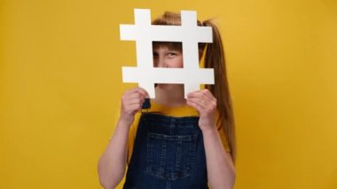 Neşeli küçük liseli kız portresi büyük beyaz hashtag sembolü ve gülümseyen çocuk blogu arkasında saklanıyor, sarı stüdyo arka planında izole edilmiş. İnternet eğilimleri, viral web içeriği