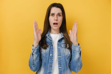 Utangaç genç esmer kadın büyük görünmez nesneyi şekillendirir, boyutunu açıklar, mavi kot ceket giyer, sarı stüdyo arka planında poz verir, büyük bir şey görünce kafası karışır. Millet, ölçüm konsepti