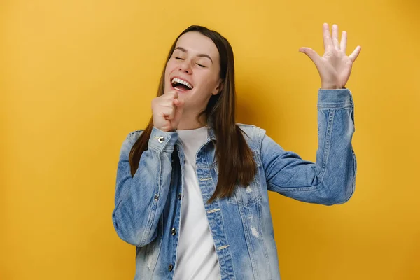 Carefree joyful millennial woman sings favorite song, holds hand as microphone near mouth, raises palm, feels relaxed and entertained, enjoys moment, wears denim jacket, isolated on yellow background