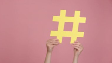 Kadın elinin altında büyük sarı hashtag sembolü, sağa sola sallanan, ünlü medya içeriğinin esrar işareti, sosyal medya pazarlaması ve blog promosyonu, pembe stüdyo arka planında izole edilmiş,