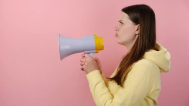 Yan görünüm üzgün beyaz kadın megafonla bağırıyor, 20 'li yaşlardaki kızgın kız öfkesini dizginlemiyor, sarı kapüşonlu giyiyor, pembe duvar arkasında izole edilmiş poz veriyor.