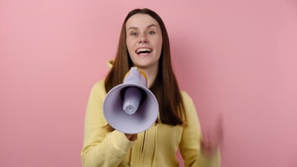 兴奋的年轻女人在扩展性扩展性的扩音器中尖叫着对着你 穿着黄色的帽衫 在粉红色的背景墙上显得孤立无援 人的情感生活方式概念 — 图库视频影像
