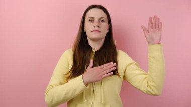 Yeminle. Dürüst, dürüst, beyaz bir kadın sadakat yemini ediyor, elini göğsüne koyuyor, avuç içi hareketler yapıyor, bir şeyler vaat ediyor, sarı kapüşonlu giyiyor, pembe stüdyo arka plan duvarından soyutlanmış.