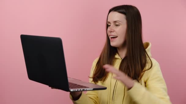 魅力的な笑顔の若い女の子20代の歳のラップトップPcでビデオ通話の挨拶 手を振って パーカーに身を包んだ ピンクのカラースタジオの背景に隔離された作業をしています 人々のライフスタイルコンセプト — ストック動画