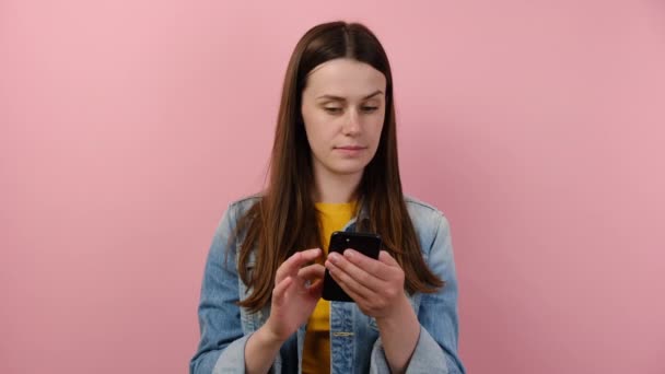 携帯電話のタイプブラウジングチャットを使用して保持夢のような普及した若い白人女性20代は ピンクの色の壁の背景スタジオに隔離された青いデニムジャケットに身を包んだ Smsを送信します アプリのコンセプト — ストック動画