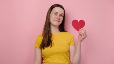 Çekici beyaz bir kadının portresi küçük kırmızı kalbi işaret ediyor, kameraya mutlu bir şekilde bakıyor, sarı tişört giyiyor, pembe stüdyo arka planında duruyor. Tatil kavramı