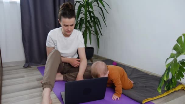 赤ちゃんを持つ若い母親は ラップトップの時計オンライントレーニングクラスの繰り返し演習の前に現代のリビングルームのマットに座っています 女性は何をするヨガの練習を選択し 自宅でラップトップのオンラインクラスを見て — ストック動画