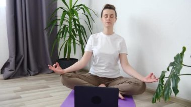 Yerde dizüstü bilgisayarla otururken meditasyon yapan ve poz veren mutlu ve çekici genç bir kadının portresi. Sakin tatlı bayan egzersiz yapıyor, nefes alıyor, yoga dersleri öğreniyor.