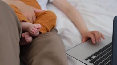Genç annenin seçici odağı dizüstü bilgisayarda klavye kullanarak küçük kız ve kullanıcı çalışanını kucağında tutuyor. Evde bebeğin yanında yatıp bilgisayar yazılımı uygulamaları teknoloji konseptiyle çevrimiçi çalışıyor.
