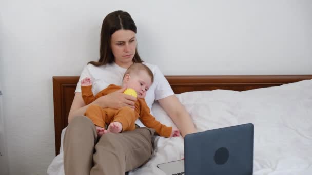 年轻母亲坐在舒适的床上工作笔记本电脑上 上网浏览电脑上的信息 亲吻可爱的小宝宝 在社交网络上与朋友交流 享受清静的周末 — 图库视频影像